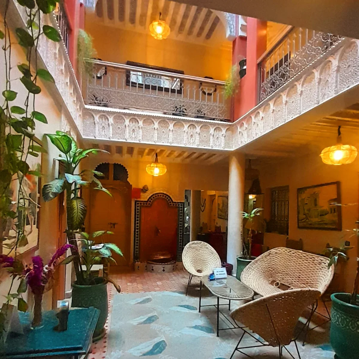 Sudut pandang interior dari courtyard tengah sebuah Riad Maroko yang elegan. Dua lantai dengan balkon bergaya mashrabiya besi tempa mengelilingi ruang terbuka. Cahaya hangat dari lampu gantung tembaga menerangi ruang tamu dengan kursi rotan dan tanaman pot besar, dengan pintu kayu tradisional di latar belakang.
