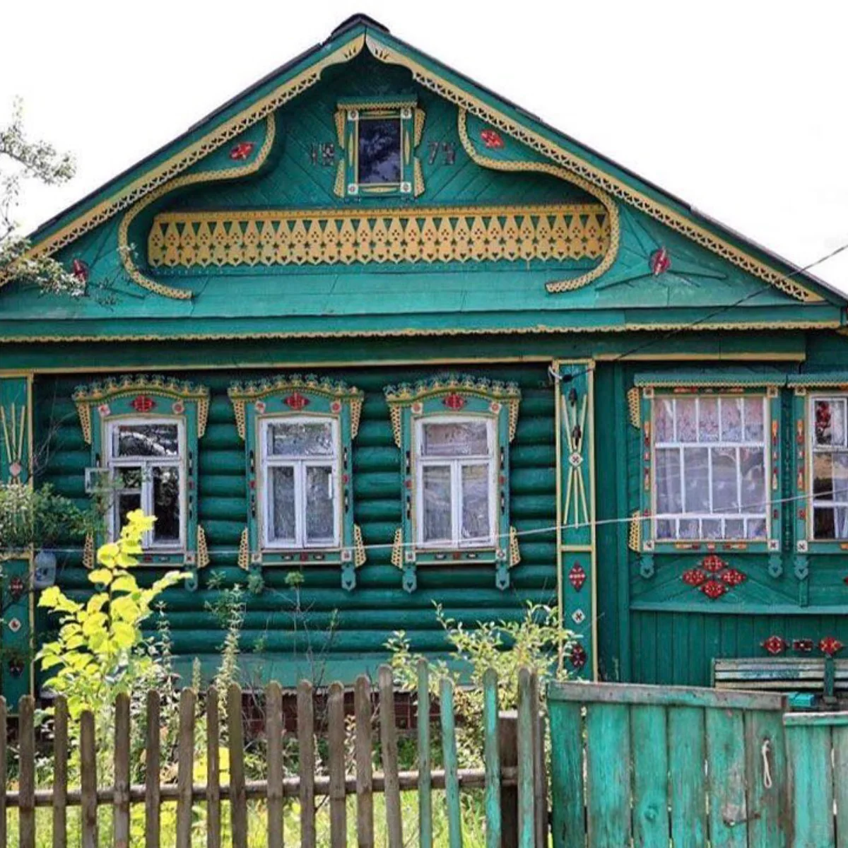 Bagian depan Izba kayu tradisional Rusia, dicat warna hijau tua yang semarak. Fasadnya dihiasi dengan ukiran kayu rumit bercat kuning dan merah di sekitar tiga jendela, pintu pagar kayu, dan atap pelana. Semak-semak hijau terlihat di bagian depan.