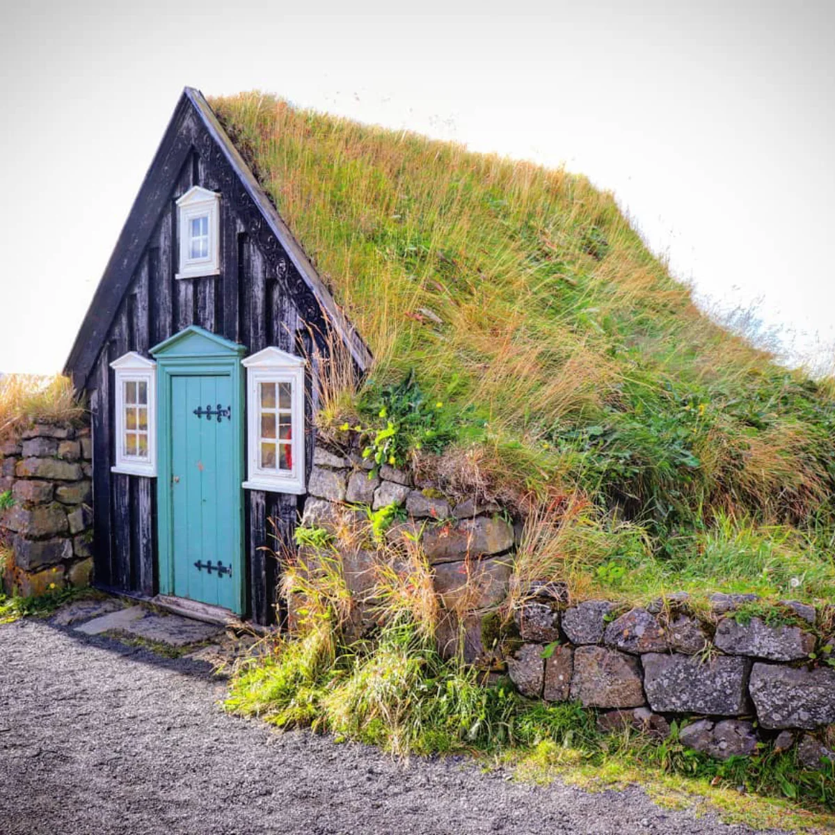 Sebuah foto dekat dari Turf House tradisional Islandia. Bagian depannya terbuat dari papan kayu hitam dengan pintu biru turquoise dan jendela putih, sedangkan atap dan sisi-sisinya ditutupi oleh rumput hijau dan tanah. Jalur kerikil mengarah ke pintu masuk.