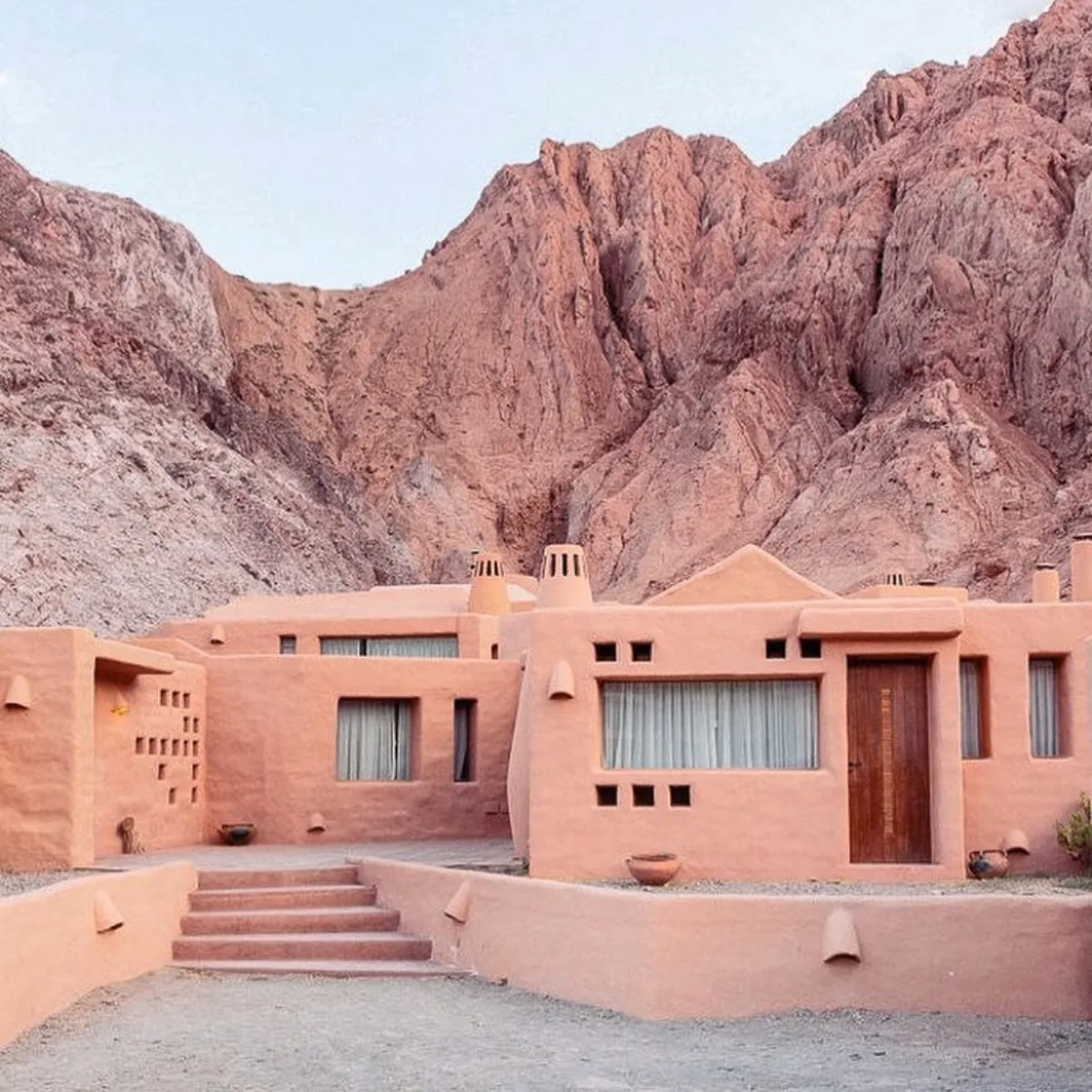 Sebuah kompleks rumah Adobe dua lantai yang dicat warna terracotta, dengan dinding luar yang tebal dan melengkung lembut serta beberapa jendela kecil. Tangga beton rendah mengarah ke teras, dengan pegunungan berbatu merah-cokelat yang besar sebagai latar belakang.