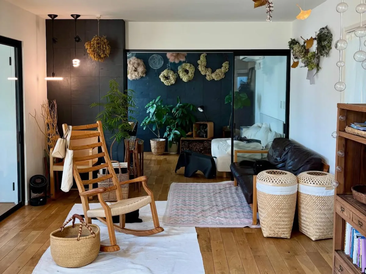 Ruang tamu dengan sentuhan natural dan rustic menampilkan kursi goyang kayu dinding aksen gelap berhias bunga kering serta keranjang anyaman untuk penyimpanan dan tanaman hias