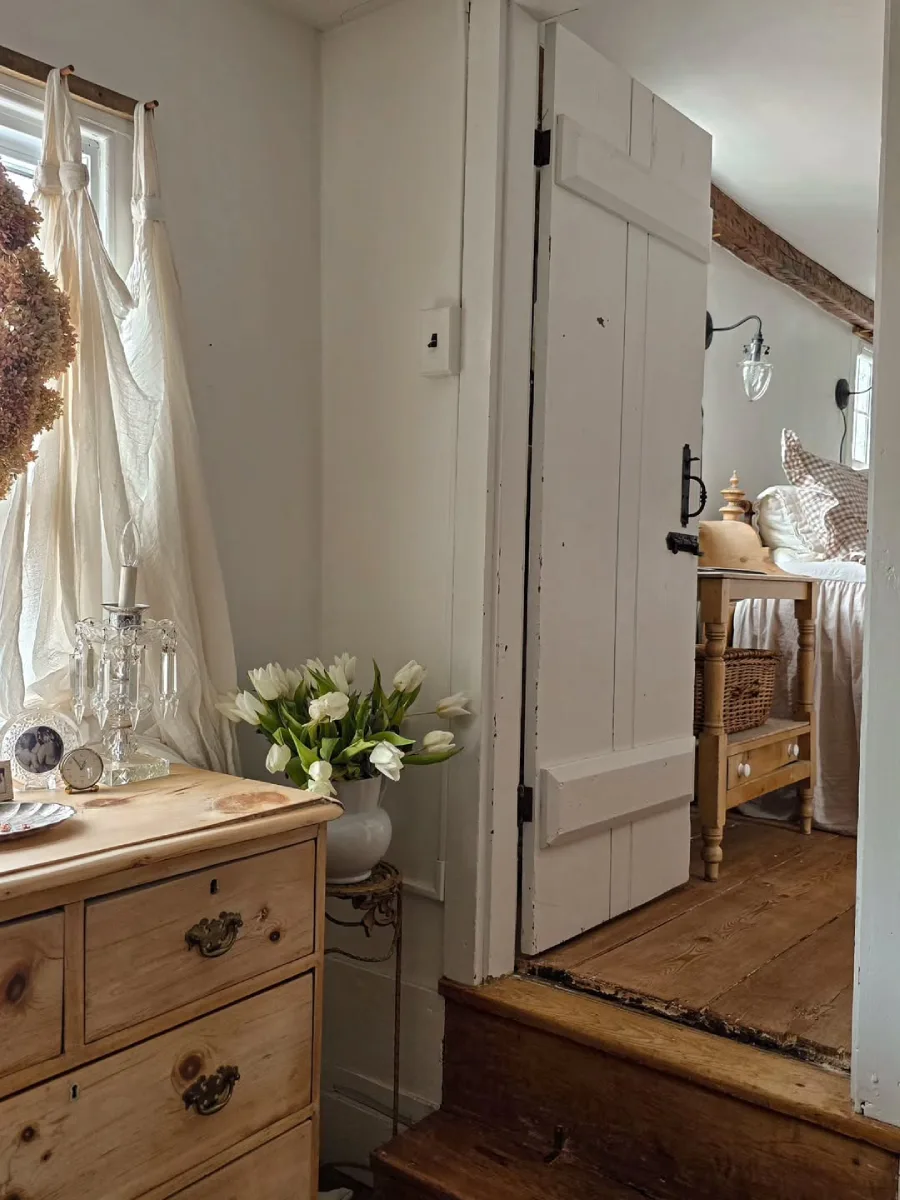 Pemandangan kamar tidur bergaya rustic melalui pintu putih terbuka yang menampilkan laci kayu alami dengan vas bunga tulip putih dan tempat lilin kristal