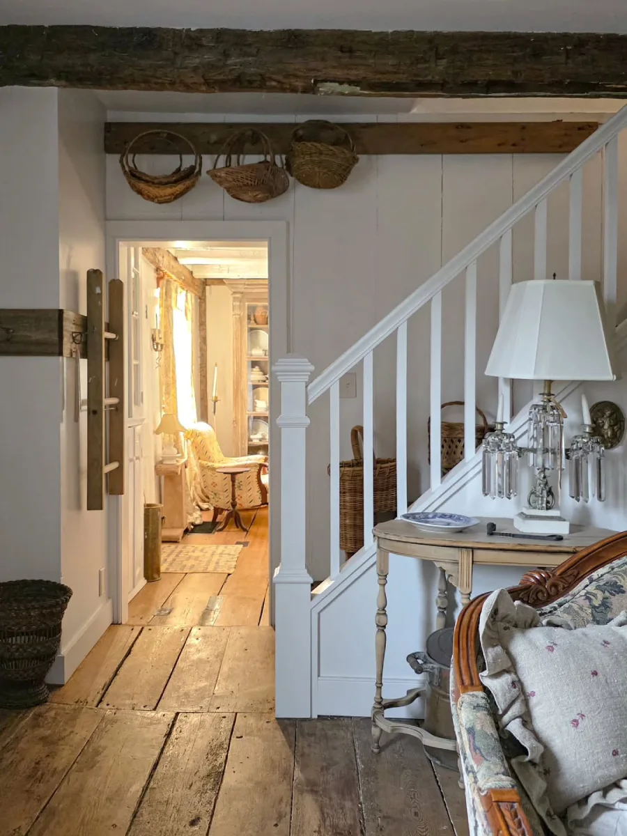 Area tangga rumah berdesain rustic dengan balok kayu ekspos di langit-langit keranjang gantung dan kursi berlengan motif floral di samping lampu vintage