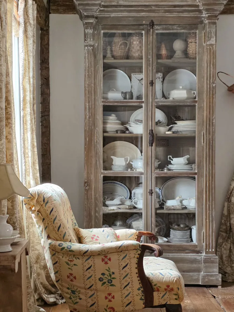 Lemari pajangan kayu antik bertekstur usang gaya rustic yang berisi koleksi piring keramik putih disandingkan dengan kursi berlengan motif bunga
