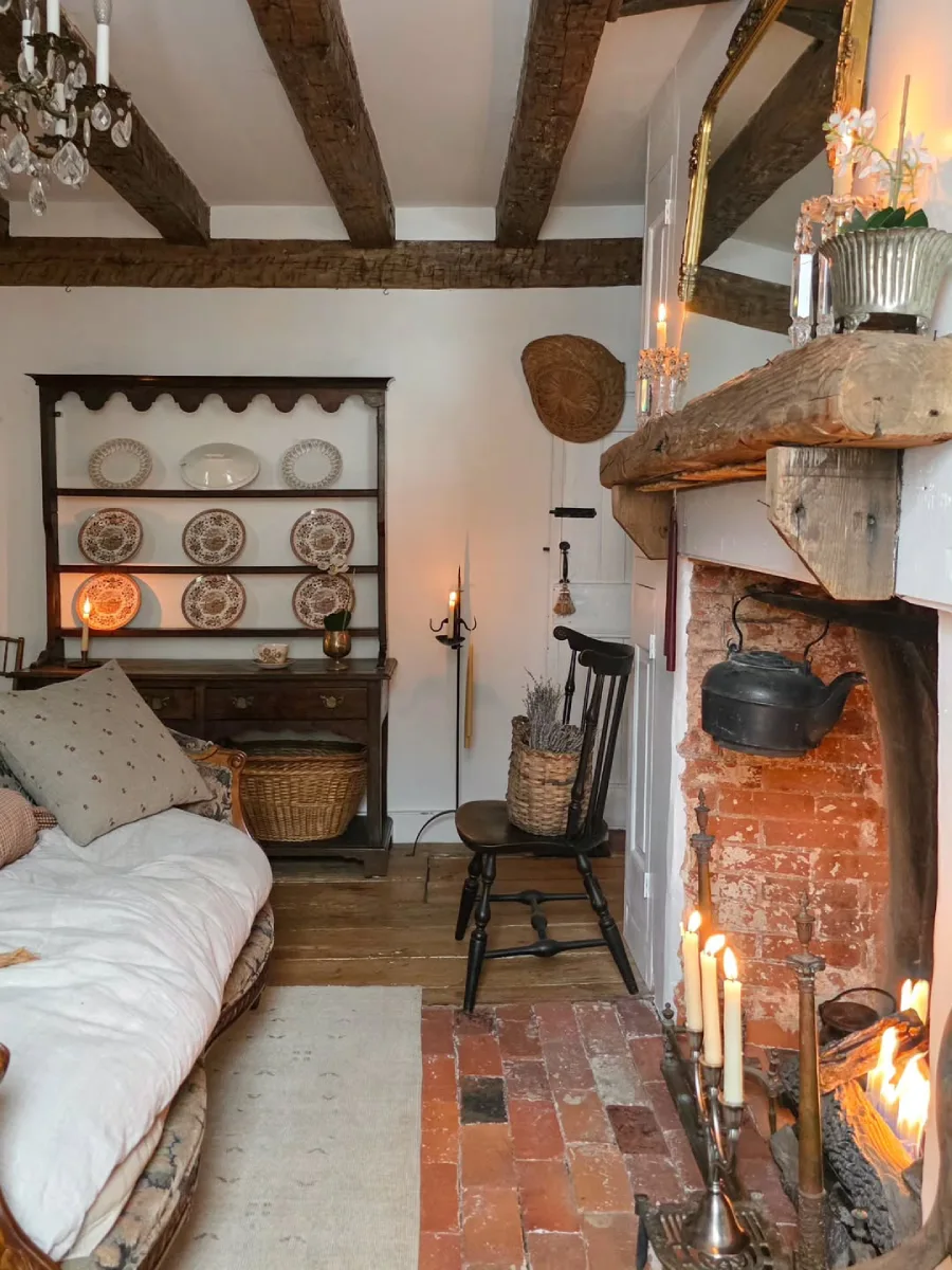 Ruang keluarga rustic yang hangat dengan perapian bata merah berisi lilin menyala rak piring kayu gelap dan sofa dengan seprai linen bertekstur