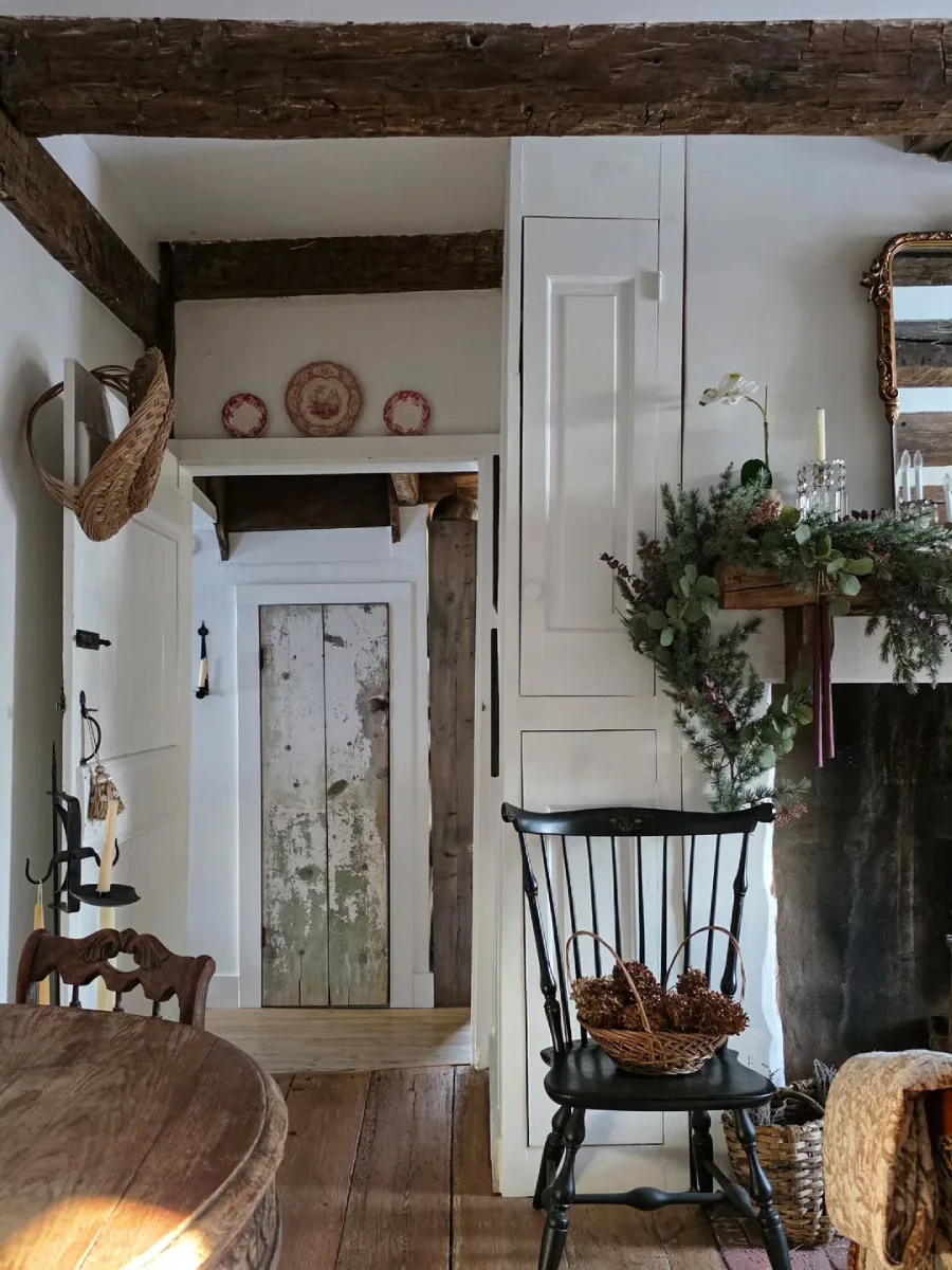 Area perapian rumah pedesaan bergaya rustic dengan hiasan garland hijau kursi kayu hitam berisi keranjang buah pinus dan balok plafon ekspos