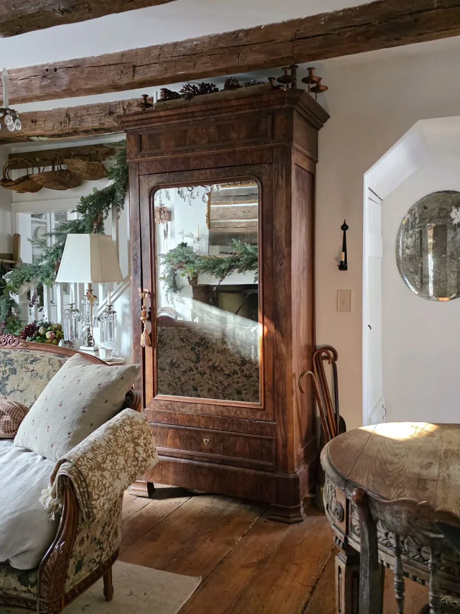 Lemari kayu antik besar dengan pintu cermin yang memantulkan ruangan bergaya rustic dilengkapi meja bundar berukir kayu dan kursi bersantai