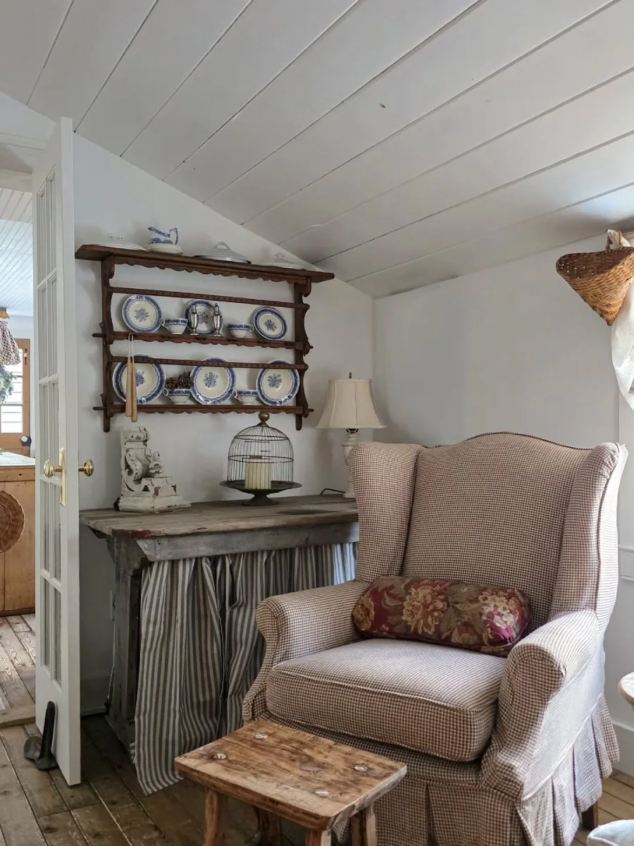 Sudut santai yang nyaman di ruangan rustic dengan kursi wingback motif kotak-kotak meja kayu usang bertirai kain dan rak dinding berisi piring keramik biru putih