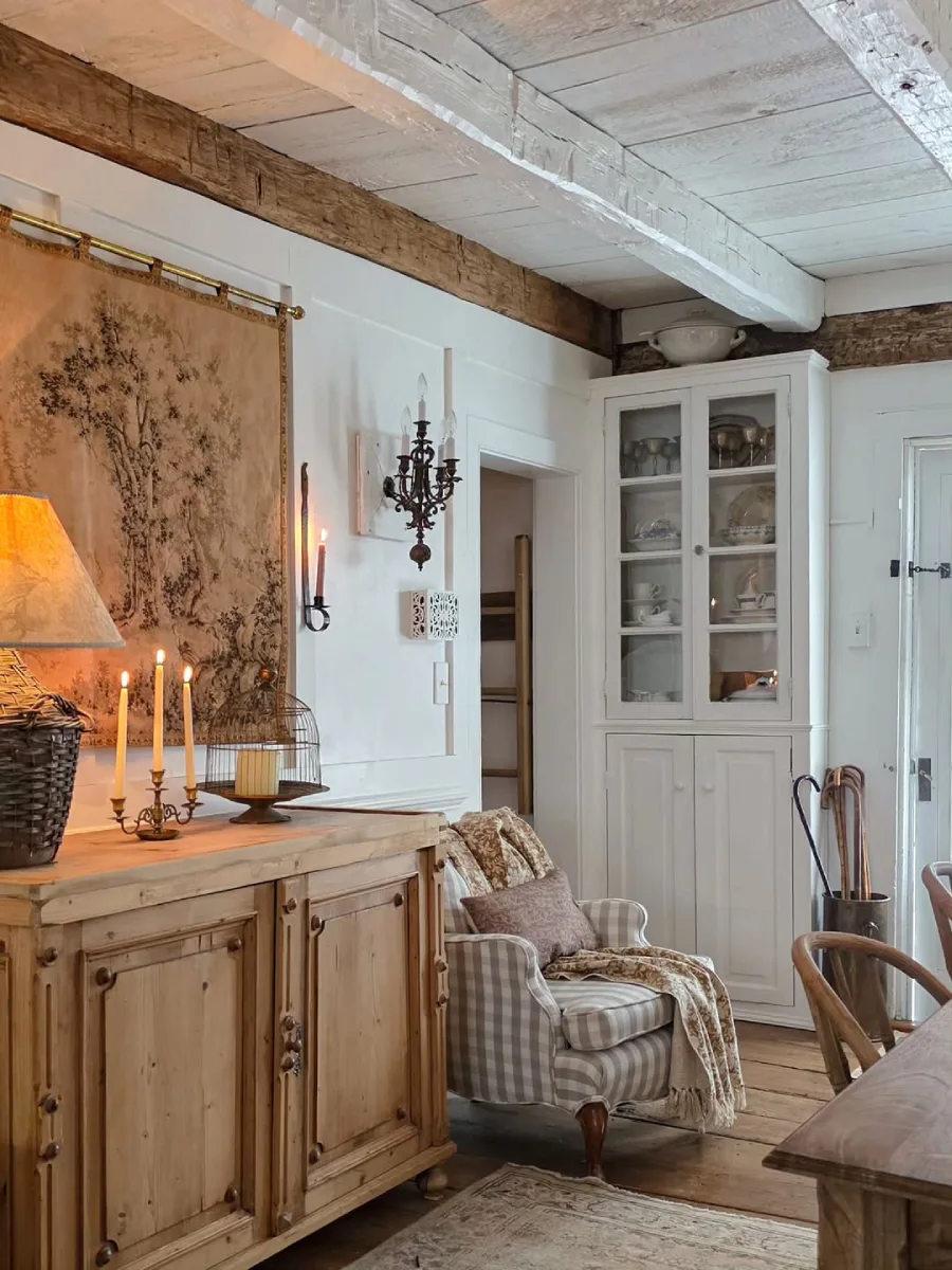 Sudut ruang makan bernuansa pedesaan dengan lemari pajangan kaca berwarna putih bufet kayu vintage kursi berlengan yang nyaman dan lantai kayu papan lebar