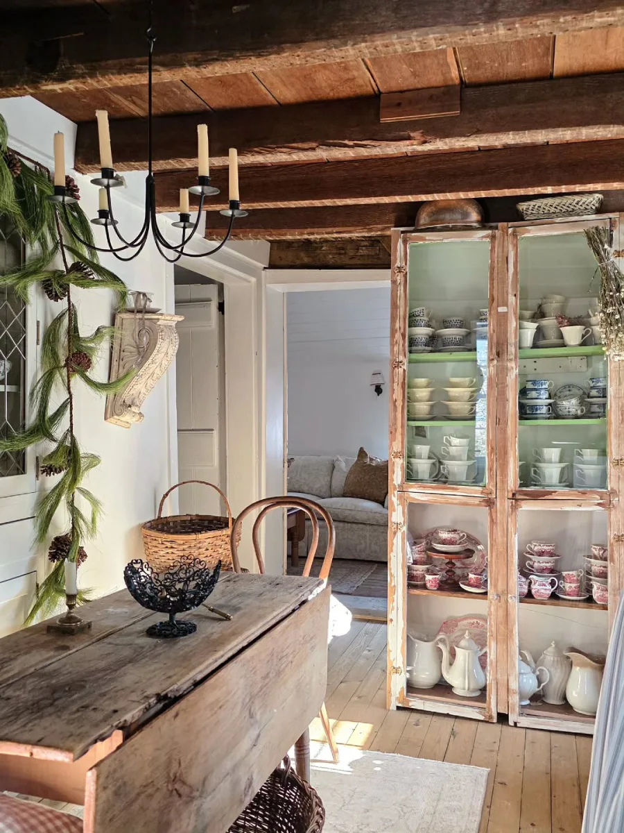 Ruang makan berdesain rustic dengan meja kayu usang, langit-langit balok terbuka, dan lemari pajangan antik berisi cangkir teh.