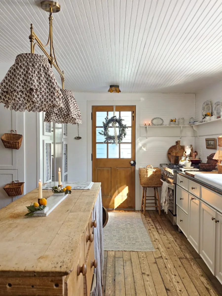 Dapur rustic cerah dengan pulau dapur kayu besar, lampu gantung kain unik, dinding panel putih, dan pintu kayu bergaya pedesaan.