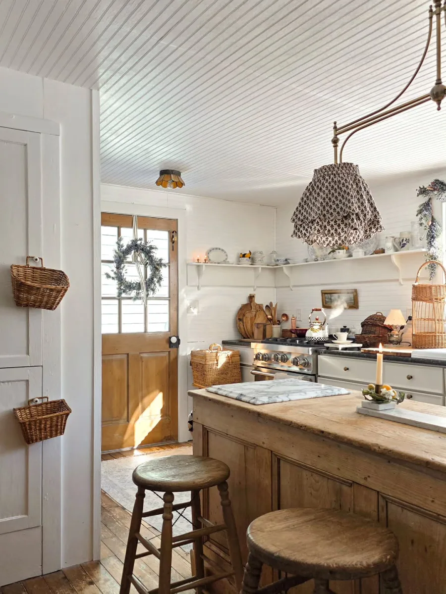 Sudut dapur bergaya rustic menampilkan meja kayu dengan bangku bulat, lantai kayu usang, dan cahaya matahari dari pintu berkarangan bunga.