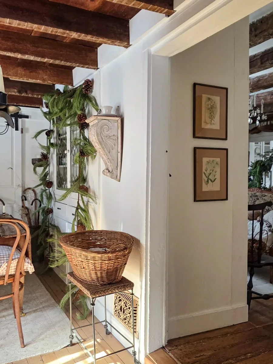 Lorong rumah rustic dengan langit-langit balok kayu, hiasan untaian daun pinus, keranjang anyaman, dan sepasang lukisan botani vintage.