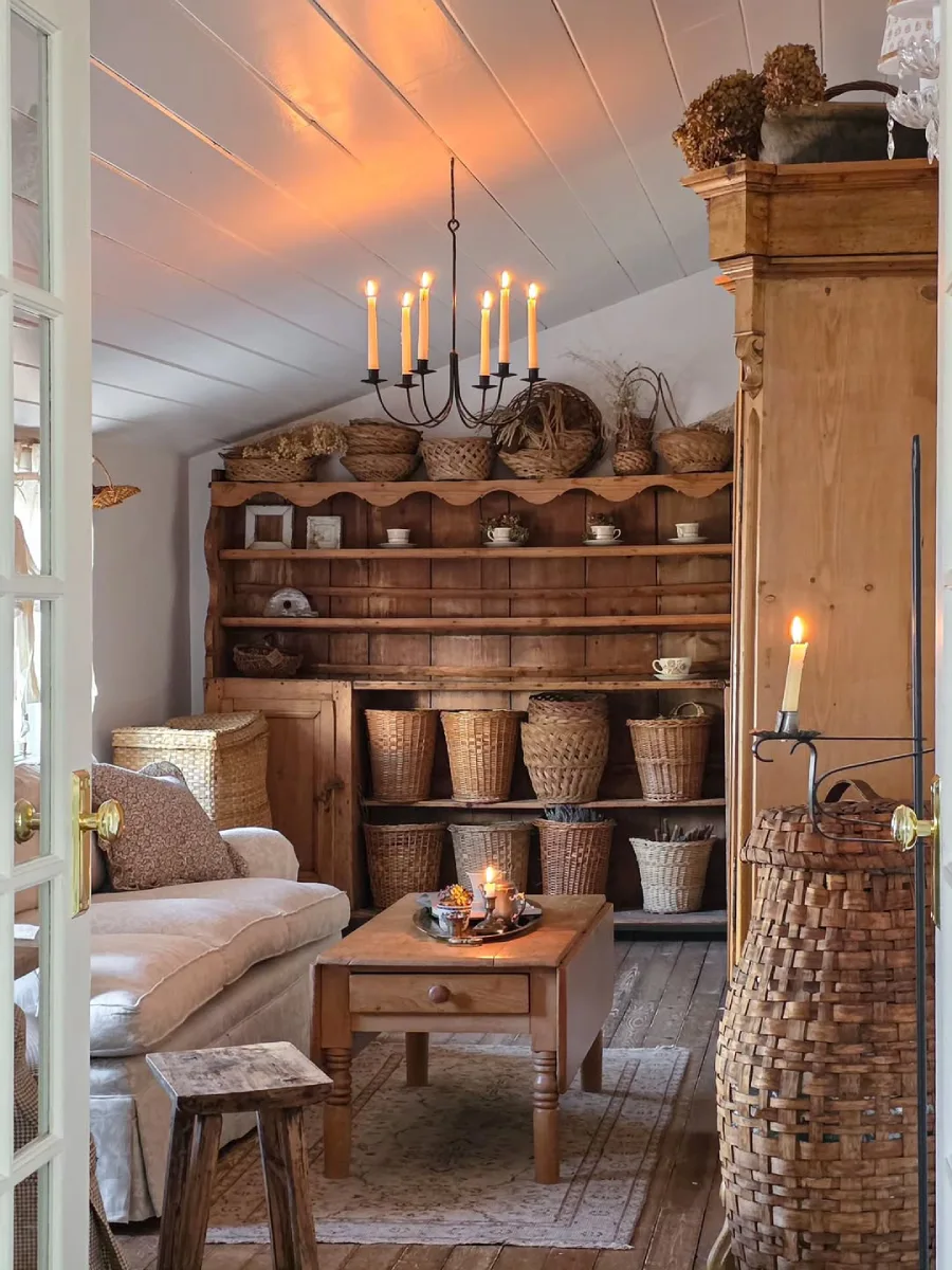 Ruang tamu rustic yang hangat dengan lemari kayu besar penuh koleksi keranjang anyaman, sofa nyaman, dan penerangan dari lampu lilin.