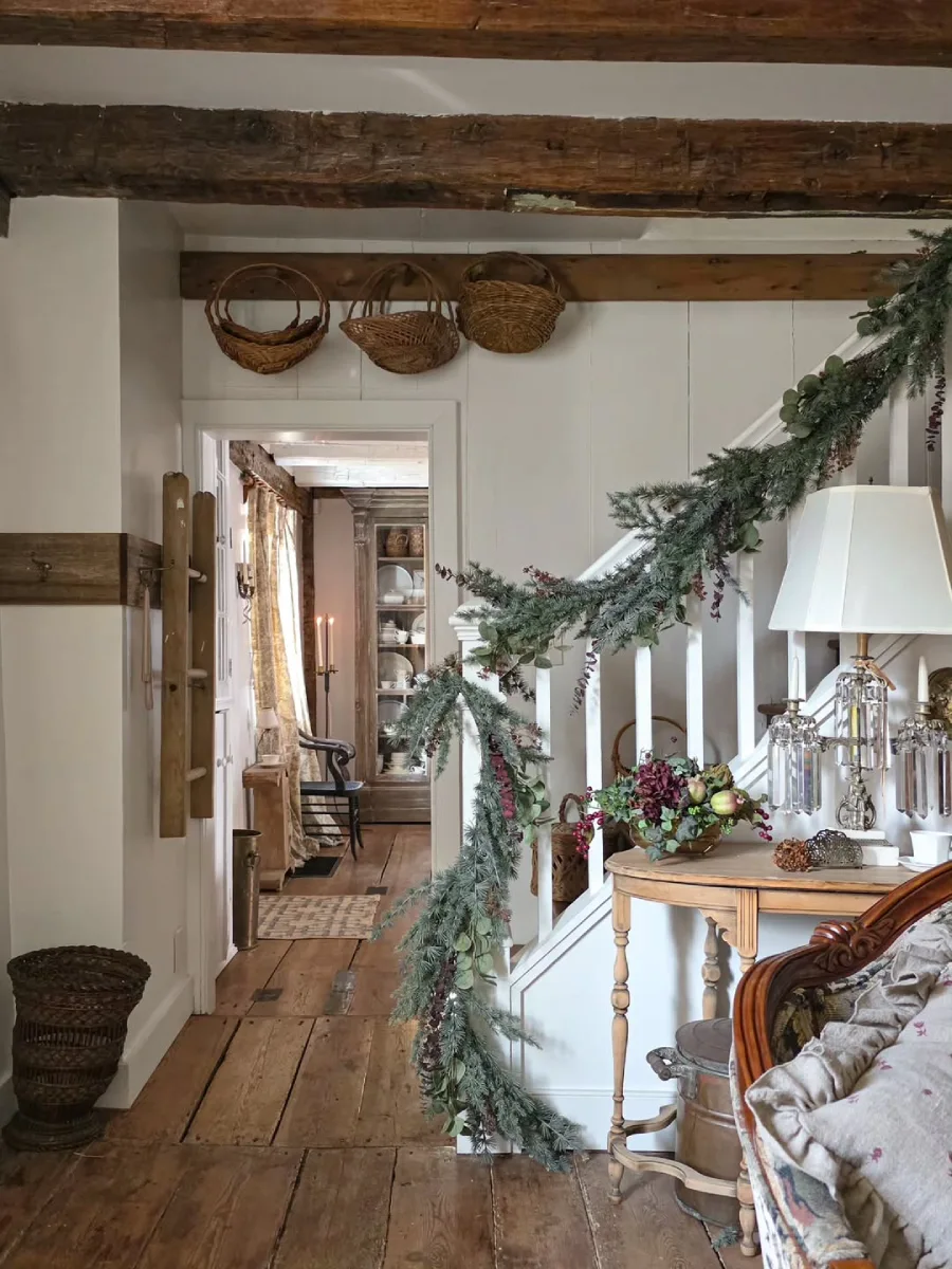 Tangga rumah bergaya rustic yang dihiasi untaian daun cemara dengan meja sudut klasik lampu kristal dan pemandangan lorong berlantai kayu usang