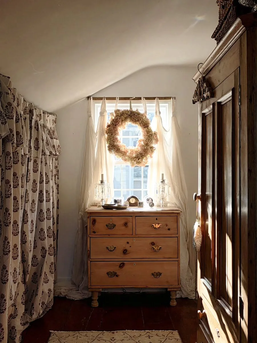 Sudut ruangan beratap miring dengan laci kayu antik berhiaskan gagang kuningan jendela berbalut tirai tipis dengan karangan bunga kering dan lemari pakaian kayu vintage