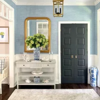 Meja konsol abu-abu di lorong masuk dengan dinding biru bertekstur cermin bingkai emas dan vas porselen biru putih berisi bunga hidrangea hijau