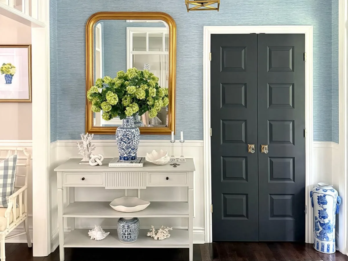 Meja konsol abu-abu di lorong masuk dengan dinding biru bertekstur cermin bingkai emas dan vas porselen biru putih berisi bunga hidrangea hijau