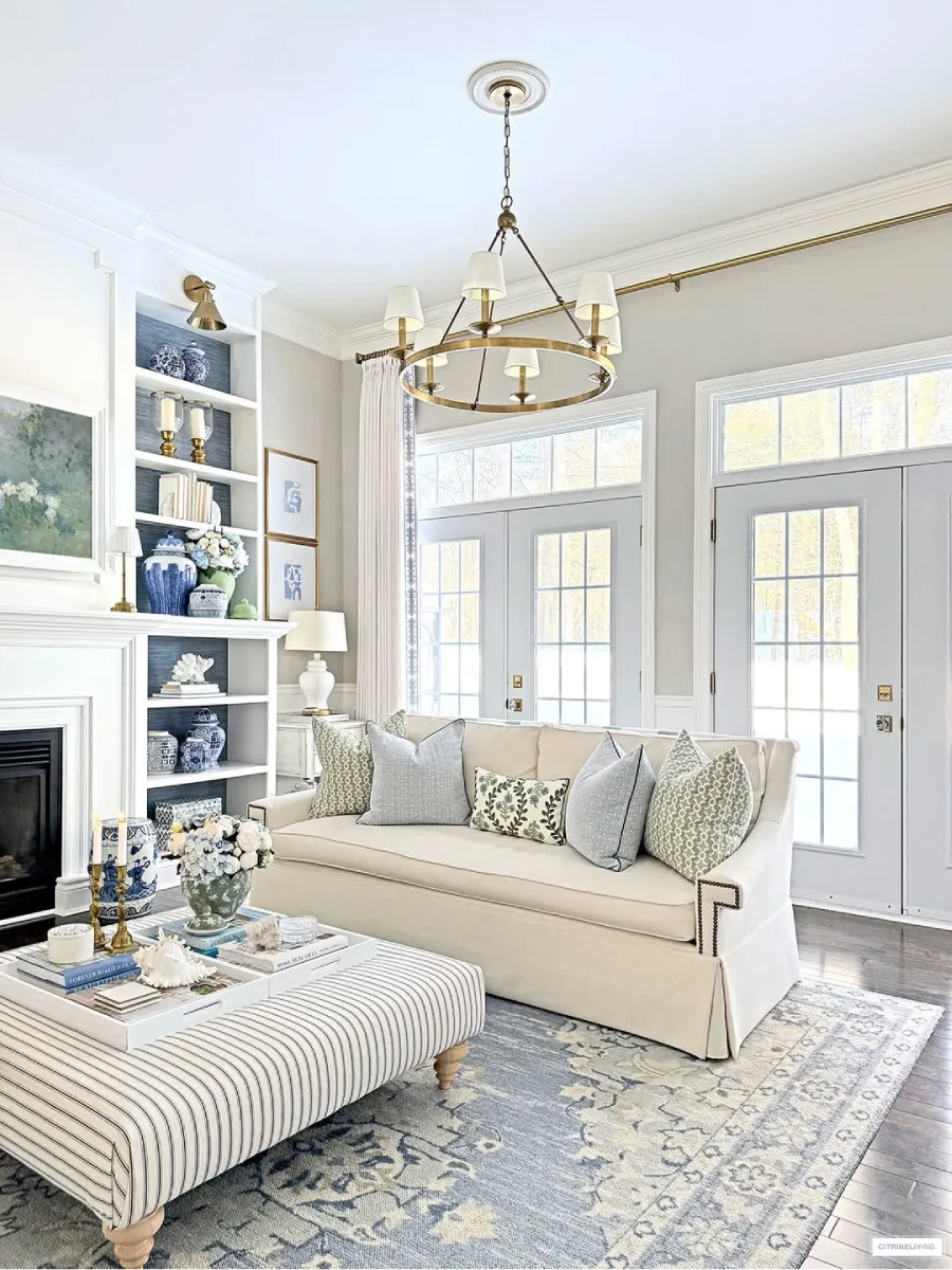 Ruang tamu transisi yang cerah dengan sofa kain krem, bantal biru-abu-abu, meja kopi ottoman bergaris biru, karpet bermotif, rak buku bercat putih dengan latar belakang biru, dan perapian. Dua pasang pintu kaca Prancis menuju luar.