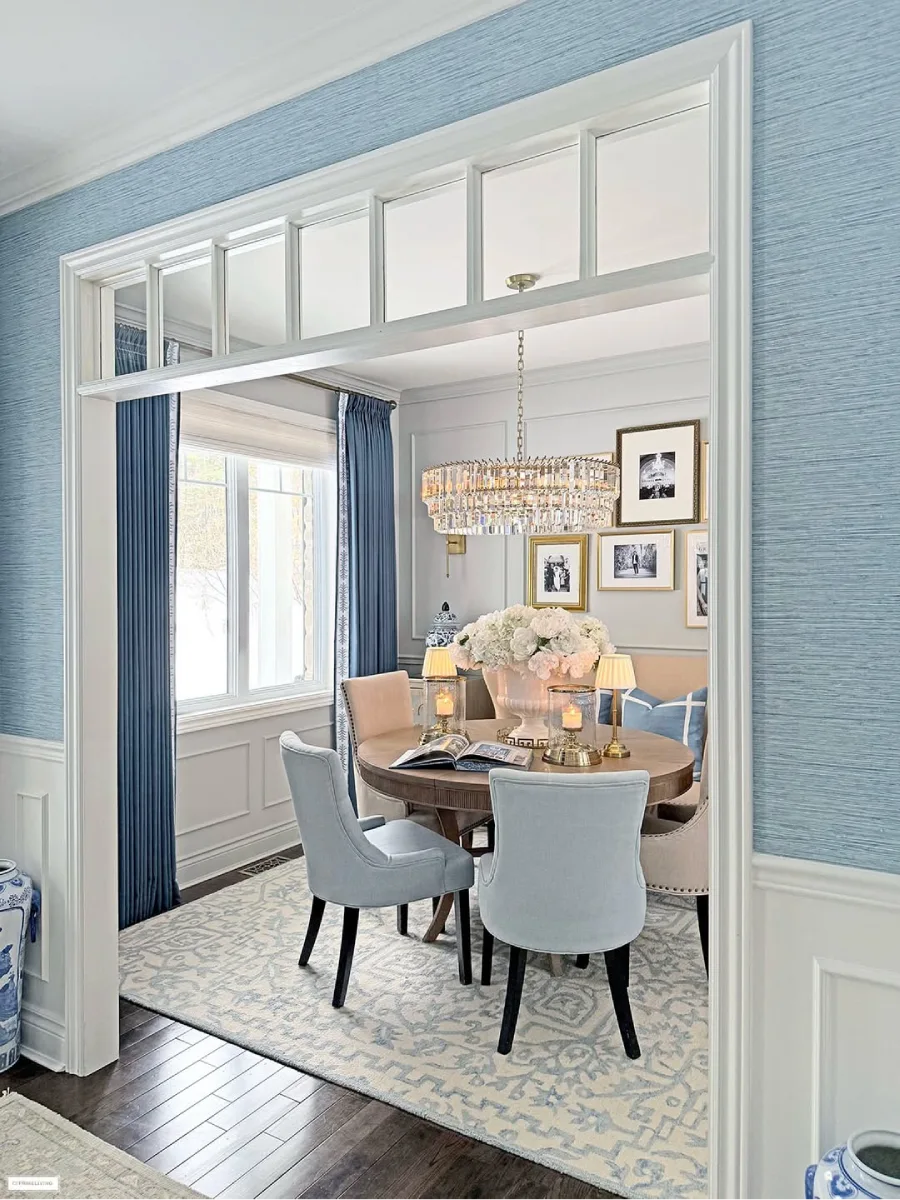 Pemandangan ruang makan dari lorong dengan wallpaper tekstur biru, menampilkan meja bundar kayu, kursi berlapis kain biru, lampu gantung kristal, dan dinding galeri foto hitam-putih.