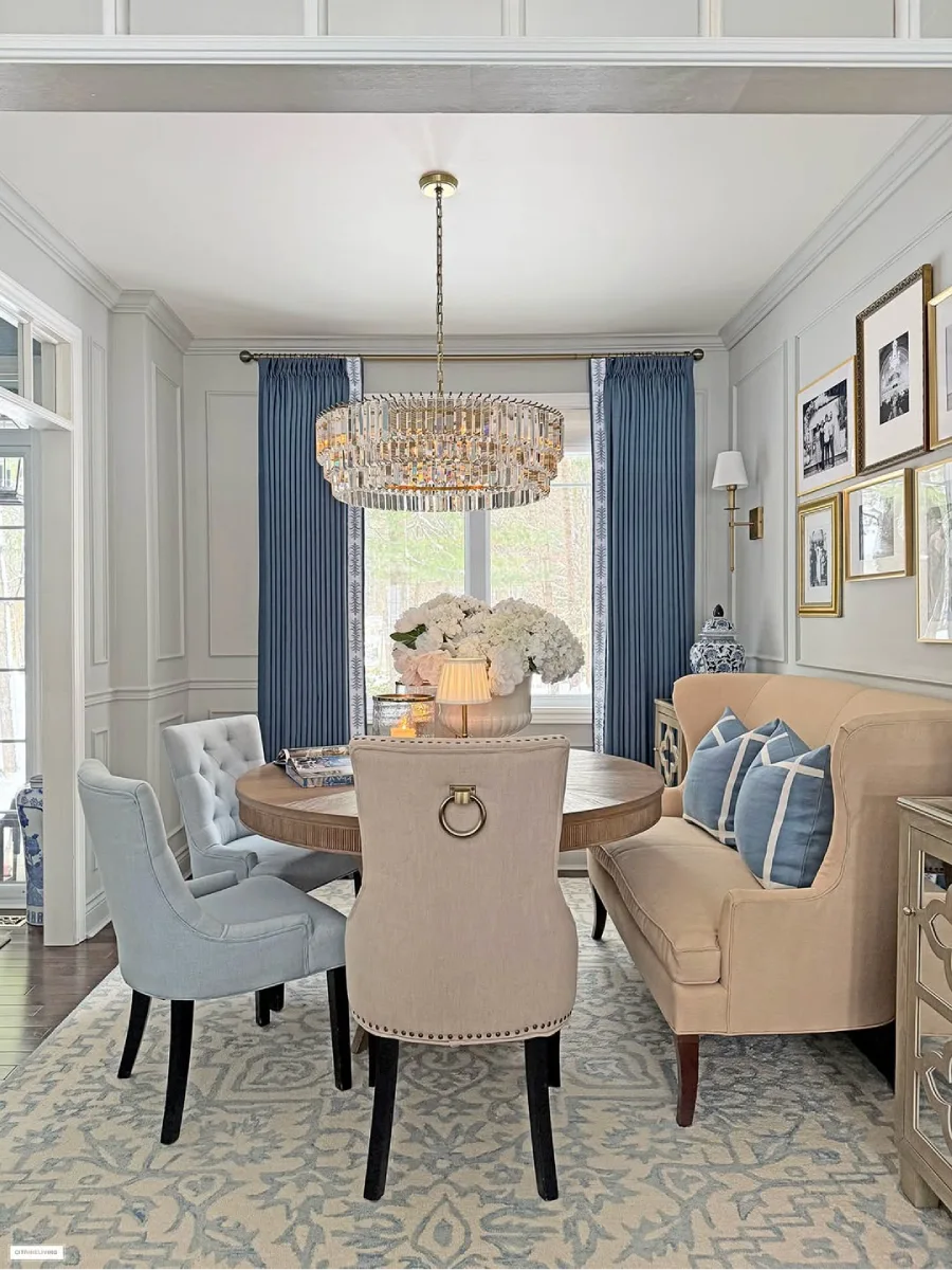 Ruang makan formal dengan wainscoting abu-abu, menampilkan meja makan bundar, bangku panjang krem dengan bantal biru, kursi makan samping biru, dan lampu gantung kristal bertingkat yang besar.