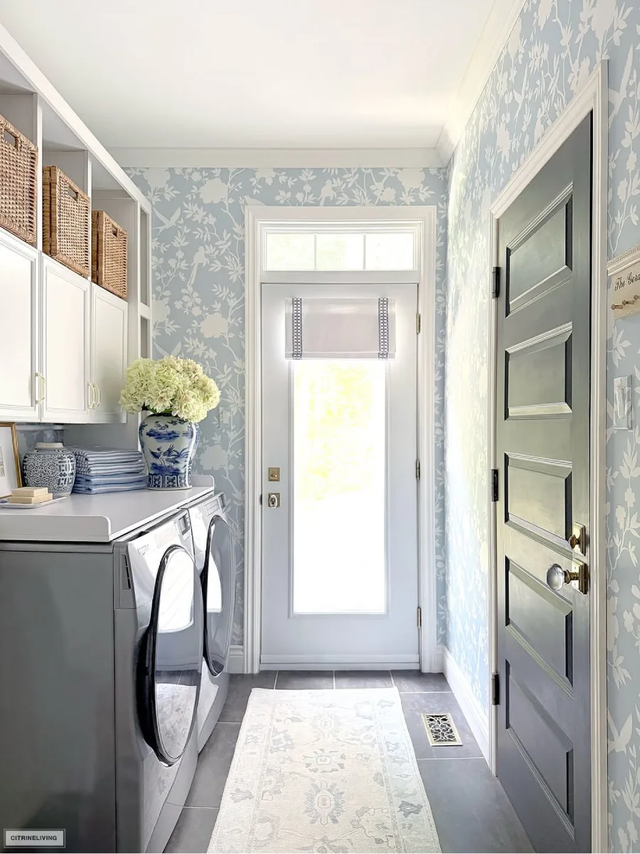 Ruang cuci dengan wallpaper floral biru muda dan putih, mesin cuci dan pengering abu-abu, lemari putih dengan keranjang anyaman, dan vas keramik biru-putih dengan bunga hidrangeas. Pintu belakang kaca dengan tirai romawi di latar belakang.