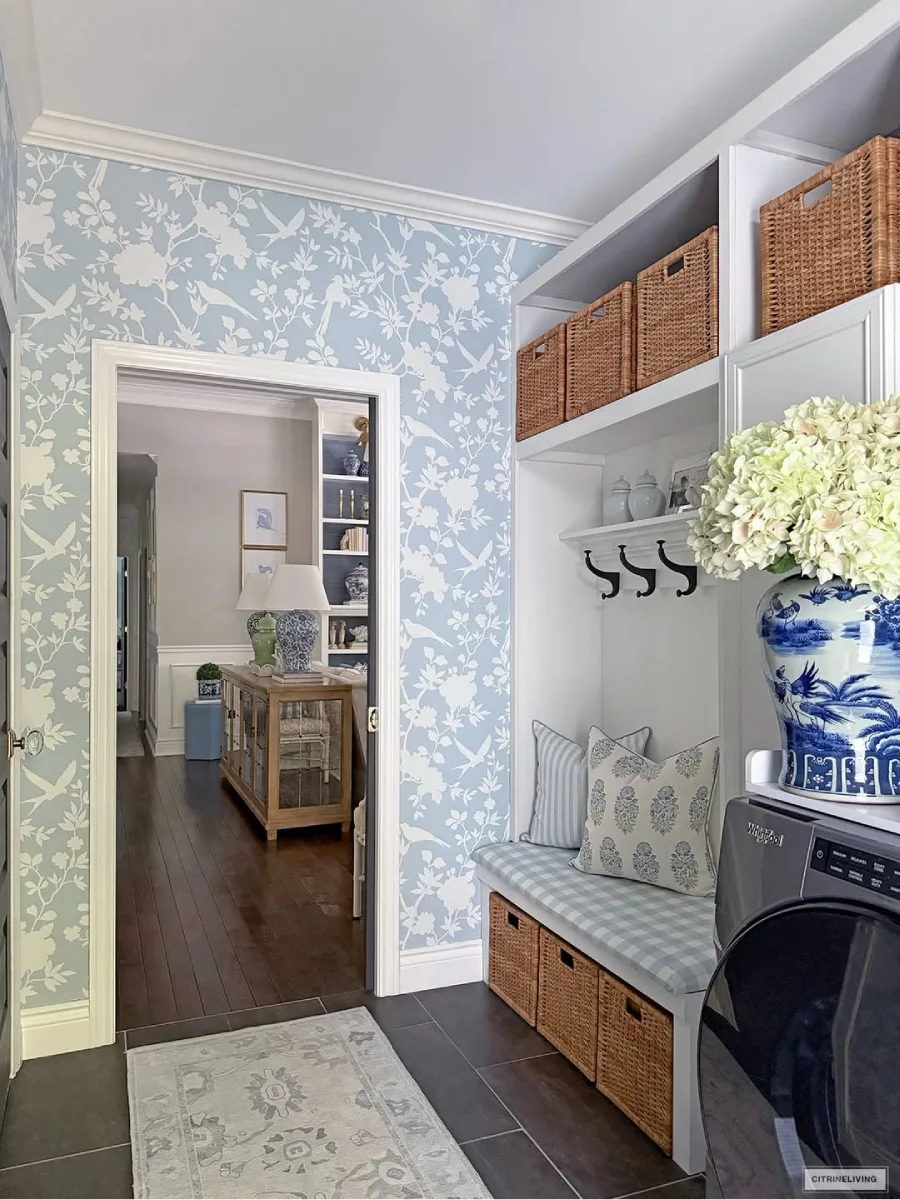 Pintu masuk atau area transisi yang menampilkan wallpaper floral biru-putih, bangku panjang berkotak-kotak dengan keranjang anyaman di bawahnya, dan pemandangan melalui pintu ke ruang tamu dengan lemari pamer kayu dan rak buku.