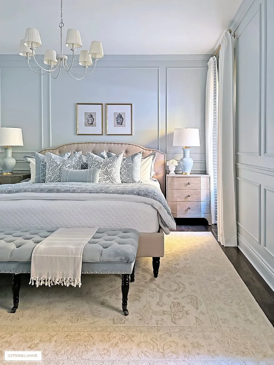 Pemandangan samping tempat tidur di kamar tidur dengan panel dinding abu-abu, tempat tidur dengan bantal bermotif biru, nakas kayu, dan bangku beludru biru di kaki tempat tidur.