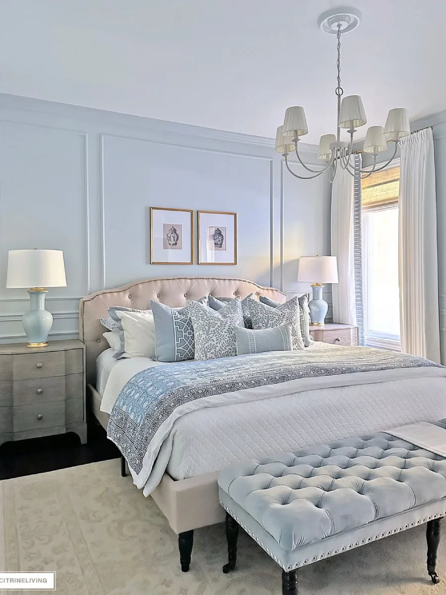 Kamar tidur utama yang elegan dengan dinding panel abu-abu, tempat tidur dengan bantal bermotif biru dan krem, lampu meja keramik biru, bangku beludru biru di kaki tempat tidur, dan lampu gantung lilin putih.