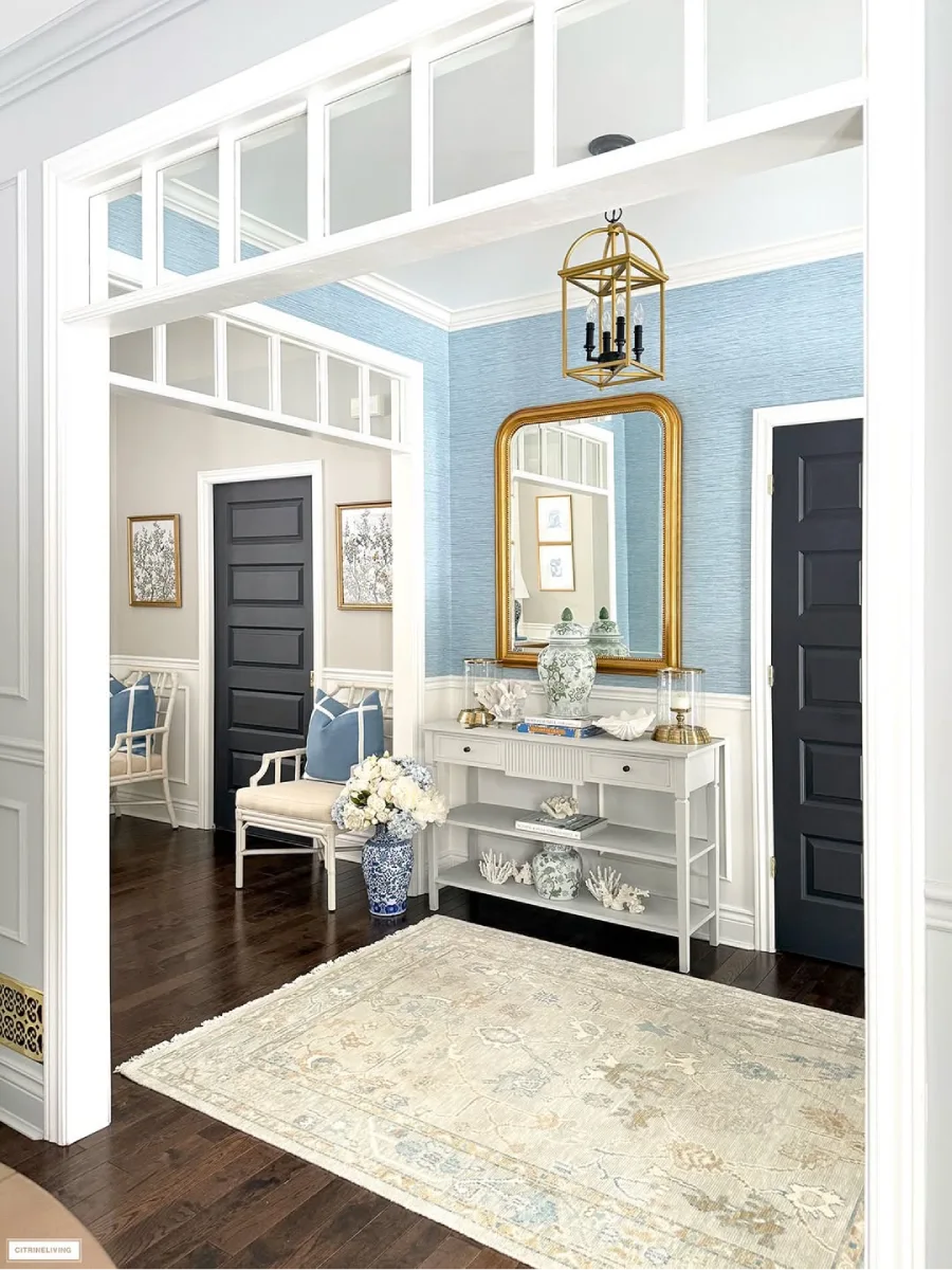 Sudut lorong rumah yang tertata rapi dengan panel dinding putih wallpaper biru bertekstur meja konsol dan pintu ganda berwarna gelap yang kontras