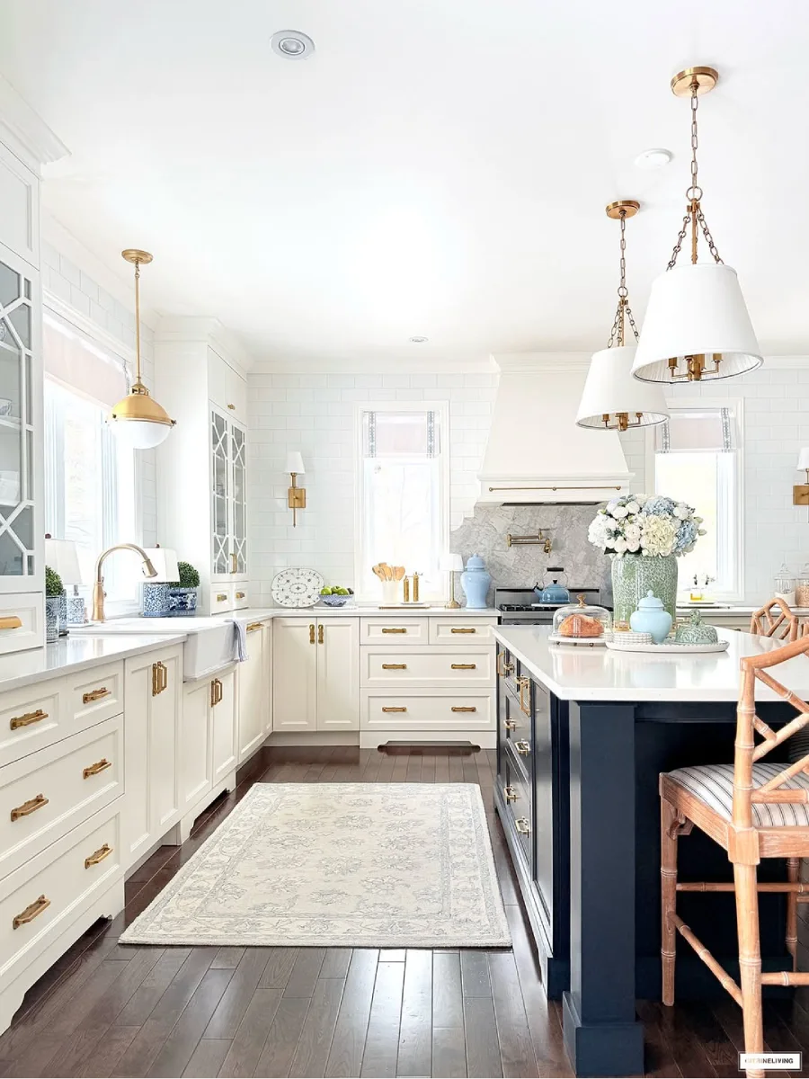 Dapur bersih dan terang dengan kabinet putih bergaya shaker perangkat keras kuningan lampu gantung emas dan kursi bar kayu di pulau dapur gelap