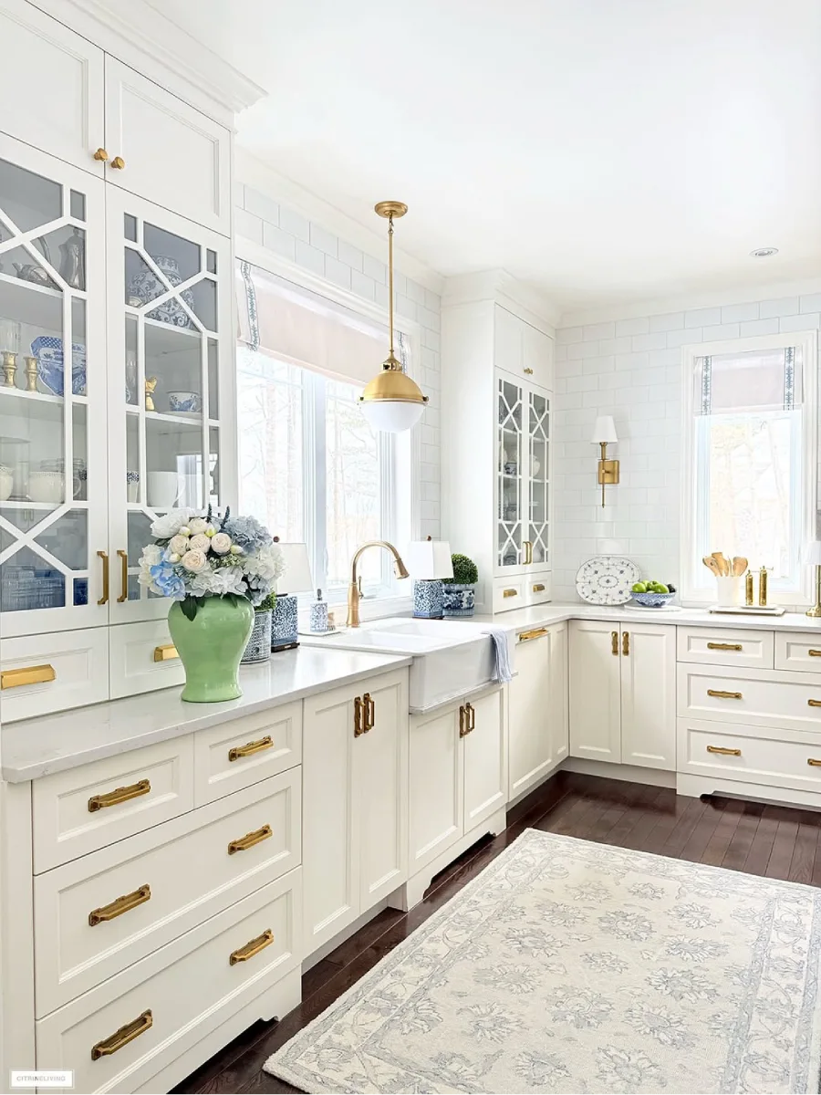 Area wastafel dapur farmhouse putih dengan keran kuningan kabinet pajangan kaca keramik biru putih dan vas hijau berisi bunga