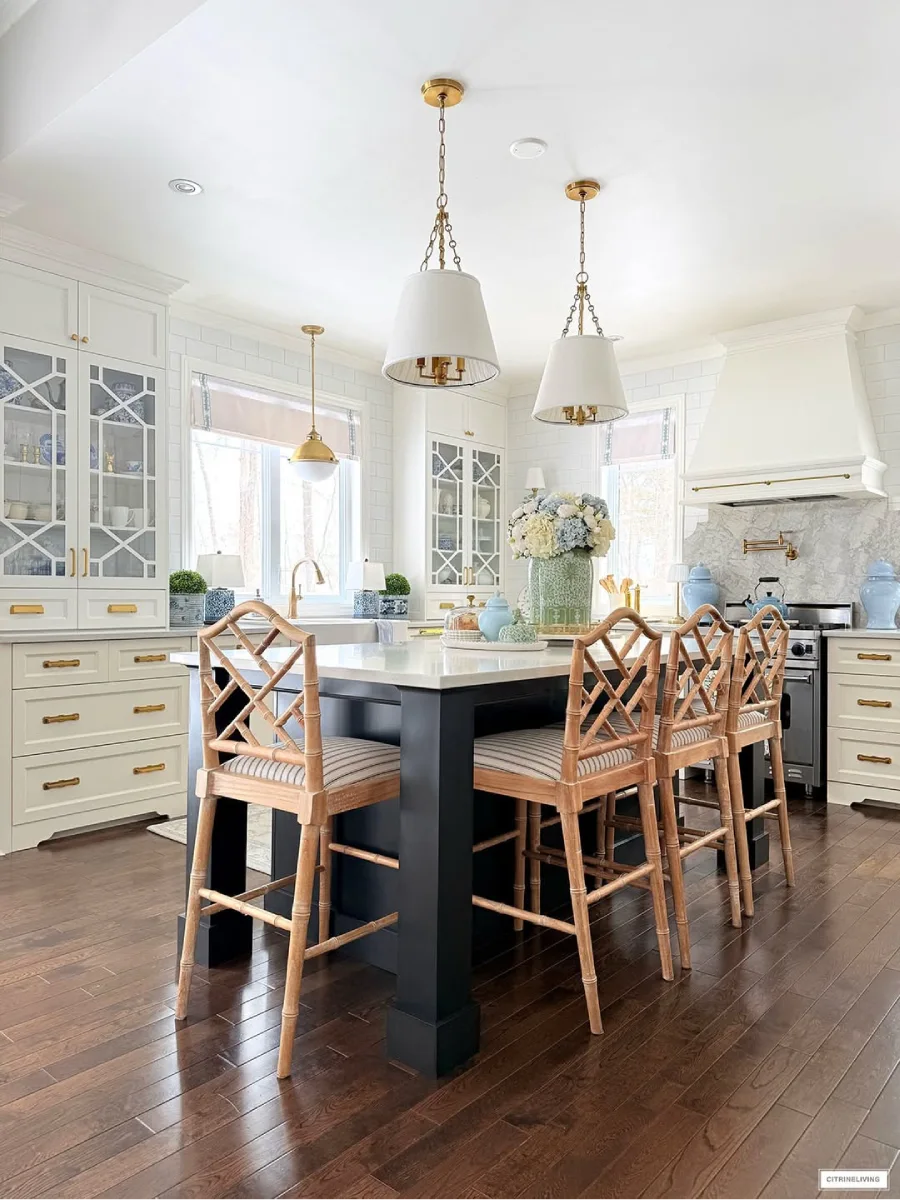 Pulau dapur berwarna gelap dengan permukaan meja putih dikelilingi kursi bar kayu dengan bantalan bergaris di bawah lampu gantung kuningan