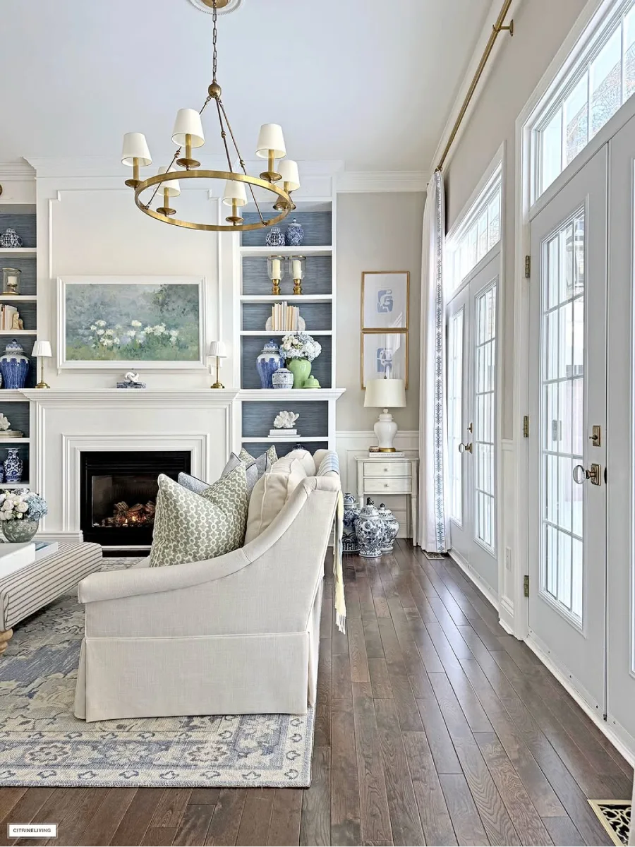 Ruang tamu yang cerah dengan sofa putih pintu kaca bergaya perancis rak buku built in berlatar biru dan dekorasi keramik di dekat perapian