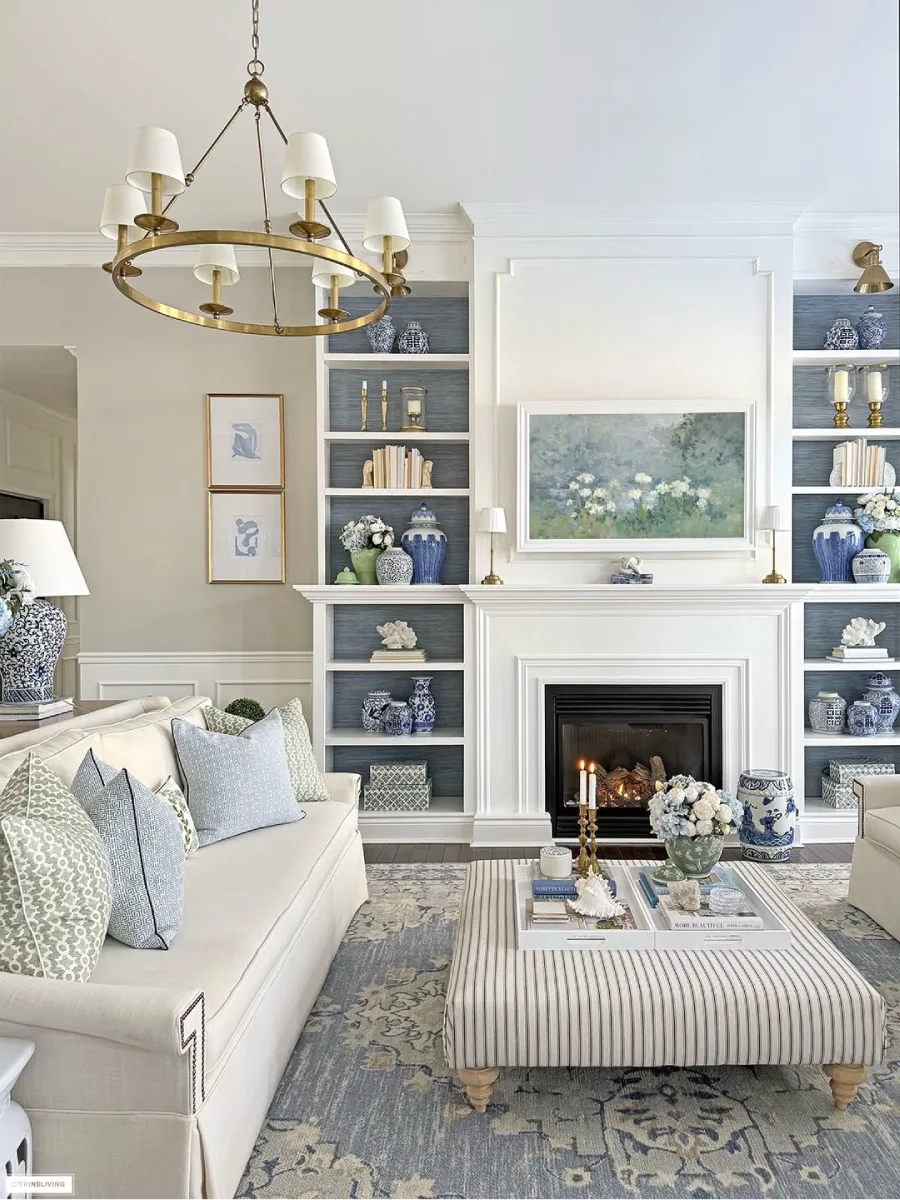 Perapian putih di ruang tamu yang diapit rak buku simetris dengan latar biru pajangan klasik dan meja kopi ottoman bergaris di tengah ruangan