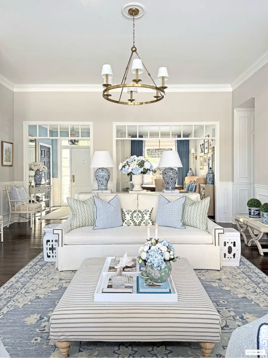 Ruang tamu bersuasana tenang dengan sofa putih bantal bermotif biru hijau lampu meja porselen besar dan pemandangan terbuka ke arah lorong pintu masuk