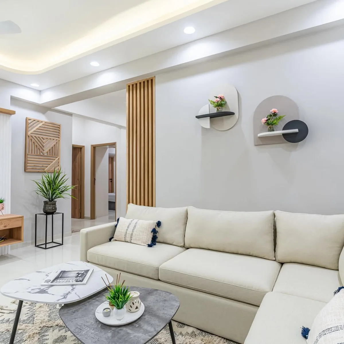 Ruang tamu modern yang cerah dengan cat tembok abu-abu muda, sofa L-shape warna krem, meja kopi unik, dan partisi kayu.