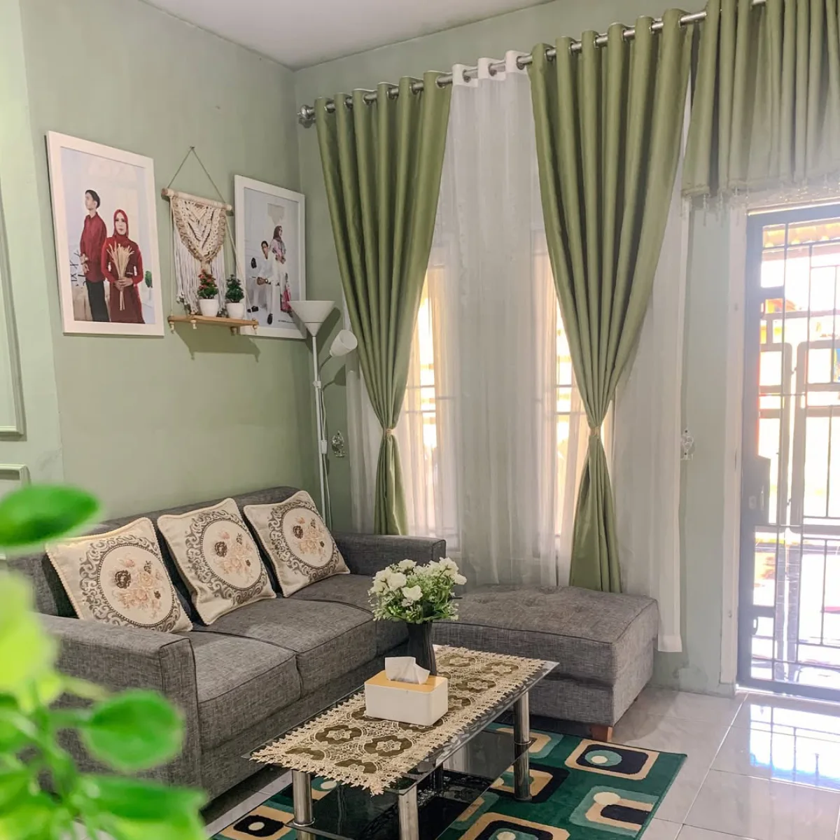 Inspirasi ruang keluarga sederhana dengan warna cat rumah hijau sage yang sejuk, gorden hijau senada, dan sofa abu-abu gelap.