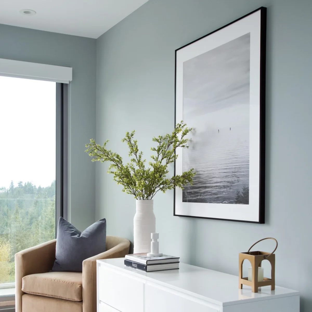 Sudut kamar tidur yang tenang dengan cat tembok biru abu-abu, kursi santai beige, lemari laci putih, dan jendela besar.