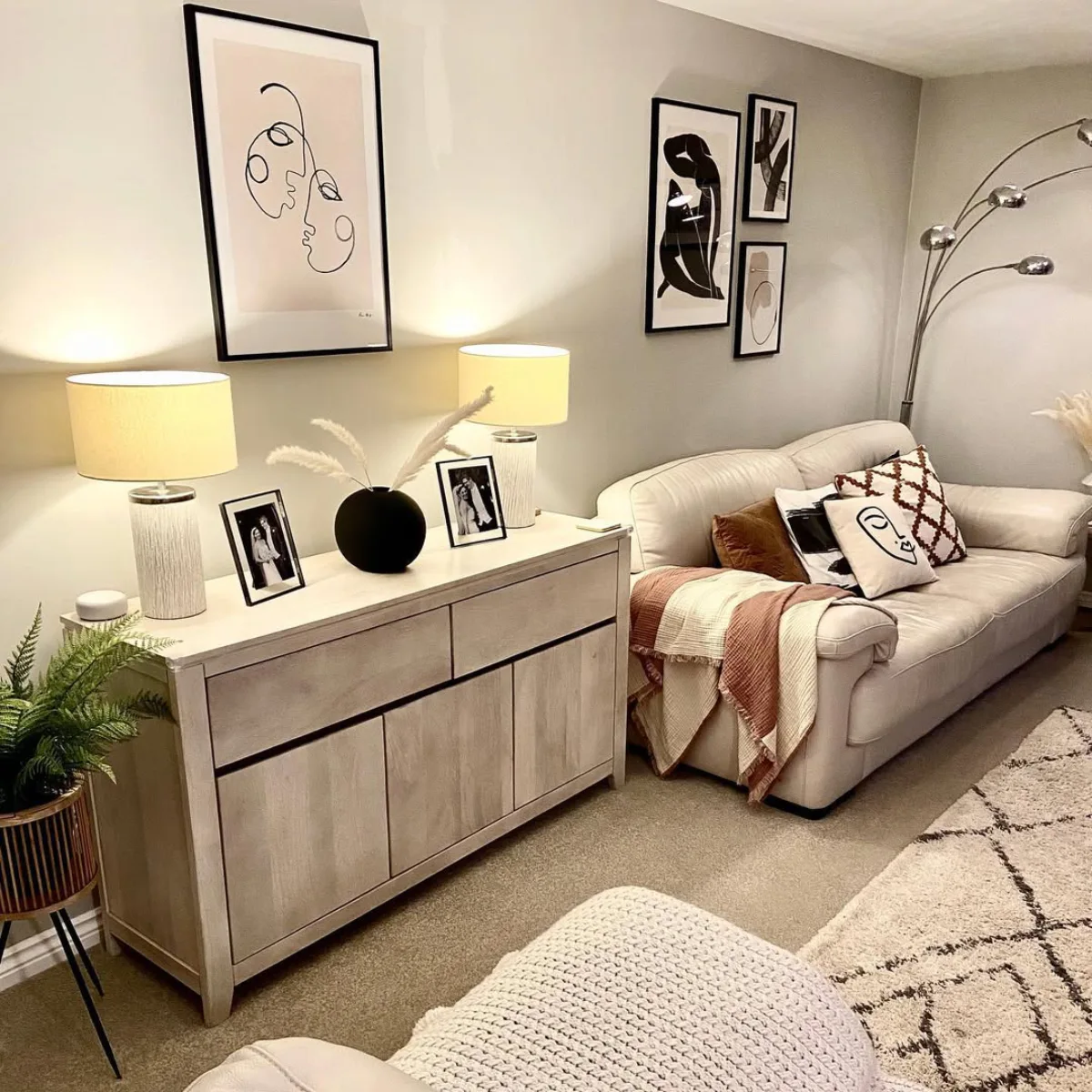 Ruang keluarga yang nyaman dengan warna cat beige abu-abu atau greige, dihiasi lampu meja hangat dan lukisan garis abstrak modern.