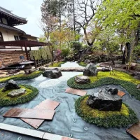 Taman Jepang dengan batu-batu besar, lumut hijau, jalur kerikil putih, dan elemen lanskap khas seperti lentera batu, serta pohon rimbun di sekelilingnya.