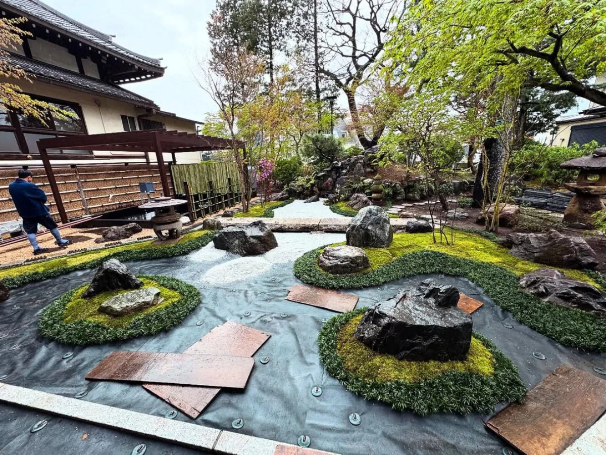 Taman Jepang dengan batu-batu besar, lumut hijau, jalur kerikil putih, dan elemen lanskap khas seperti lentera batu, serta pohon rimbun di sekelilingnya.