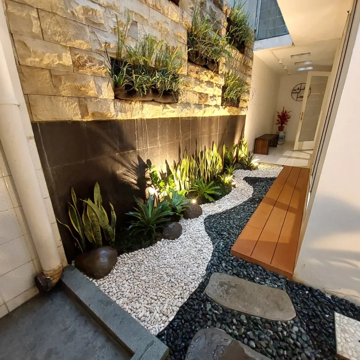 Taman kering estetik dengan kombinasi batu koral hitam putih pola gelombang, dinding batu alam, dan pencahayaan lampu sorot warm light.