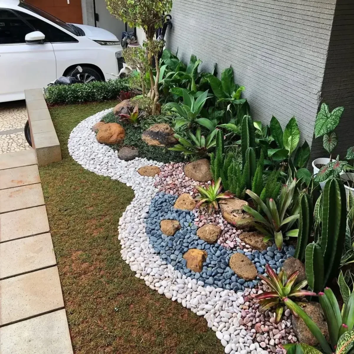 Taman kering depan rumah sebelah carport dengan desain lanskap organik melengkung, memadukan rumput jepang, batu koral hias, dan tanaman kaktus vertikal.
