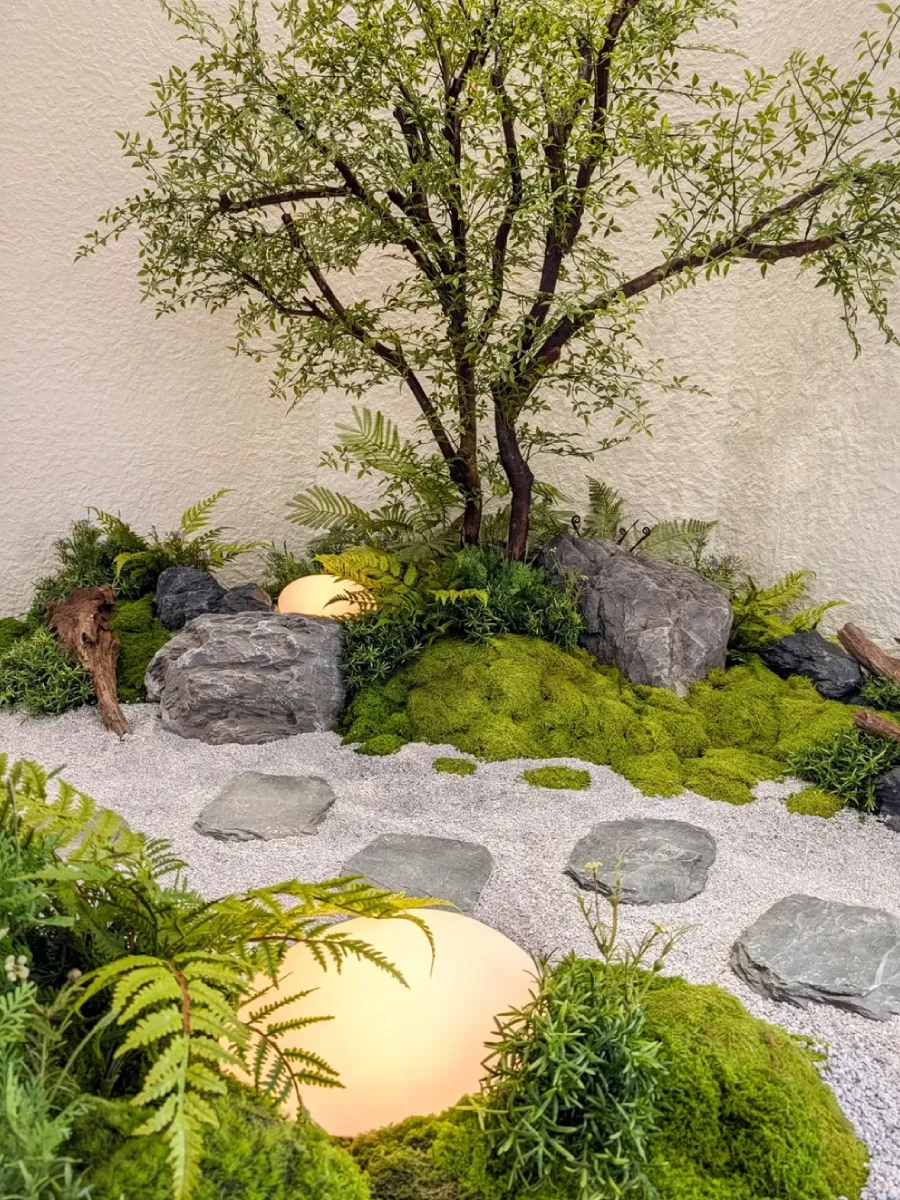 Close-up taman kering gaya Zen indoor dengan hamparan lumut hijau, ornamen batu alam, tanaman pakis, dan lampu taman bulat (ball light) yang estetik.