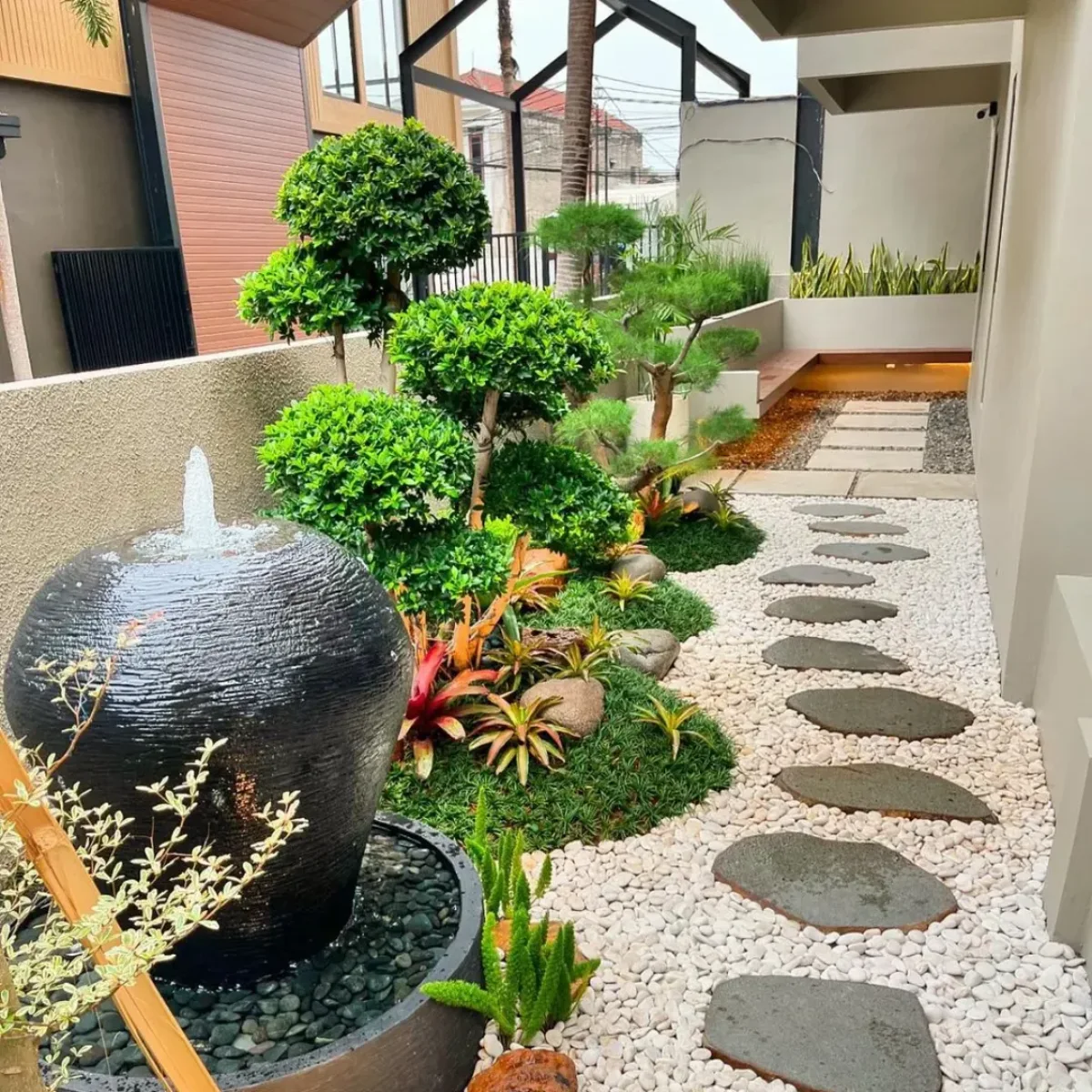 Inspirasi taman kering samping rumah mewah dengan air mancur gentong teraso hitam, pohon bonsai pangkas awan (cloud pruning), dan jalan setapak batu alam di atas koral putih.