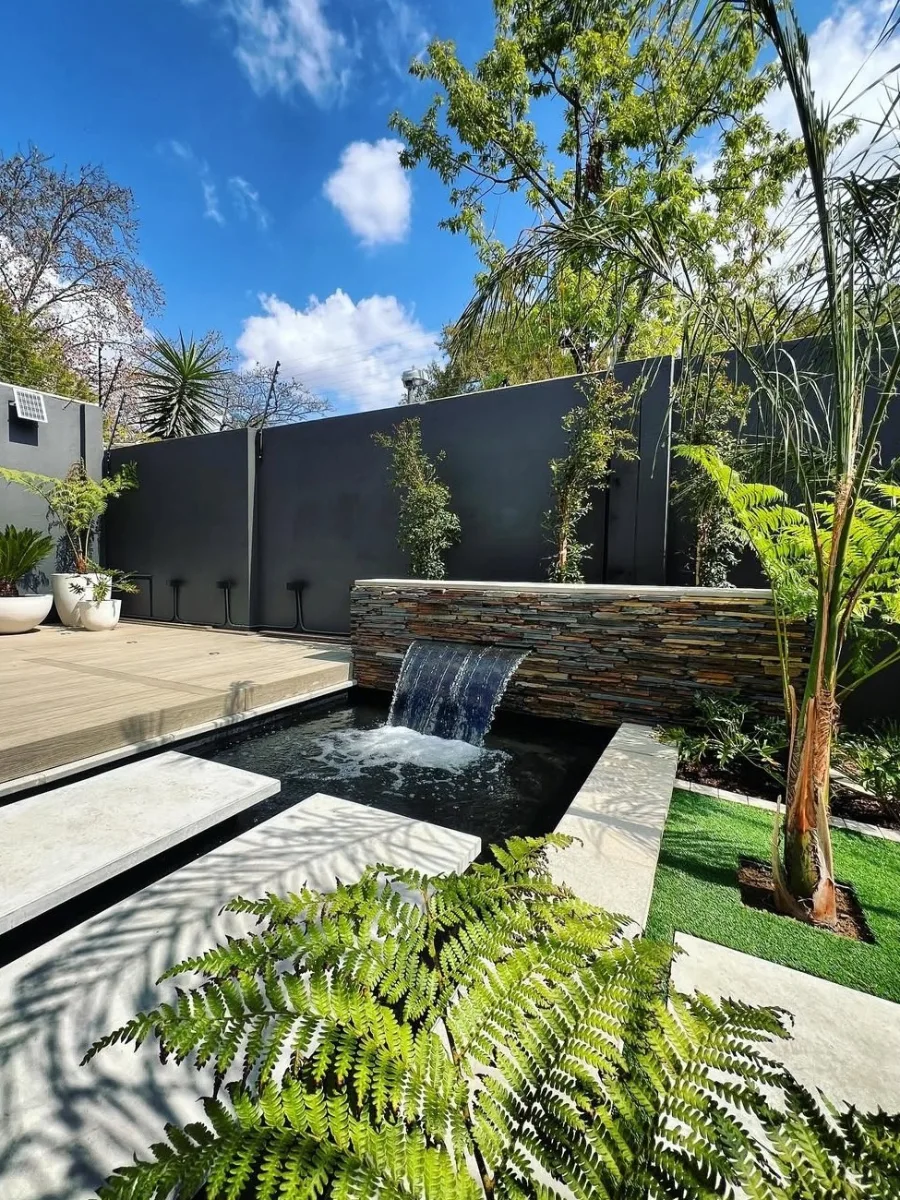 Fitur air terjun modern pada dinding batu alam dengan kolam minimalis, dilengkapi pijakan beton melayang (floating steps) dan taman rumput sintetis.