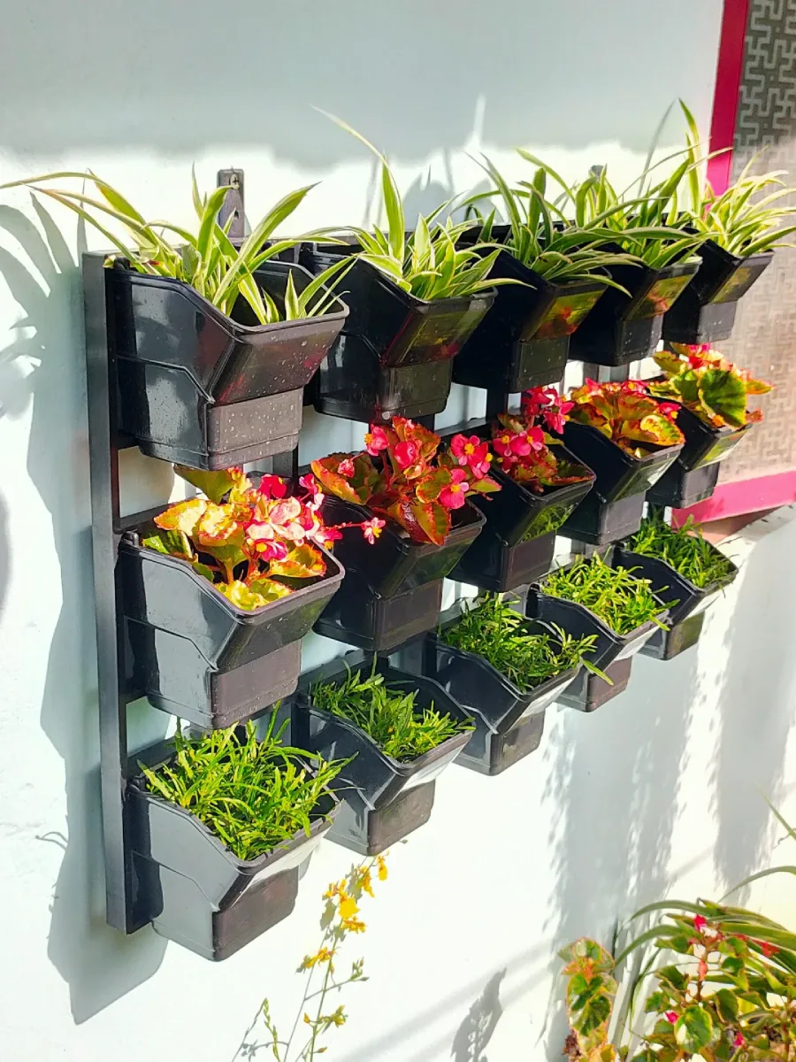 Susunan pot plastik hitam bertingkat di dinding luar, ditanami kombinasi tanaman hias hijau dan bunga warna-warni.