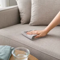 Tangan seseorang sedang mengusap permukaan sofa kain berwarna abu-abu muda menggunakan lap basah, dengan botol semprot, sikat kecil, dan mangkuk air yang tersedia di atas meja kayu di dekatnya.