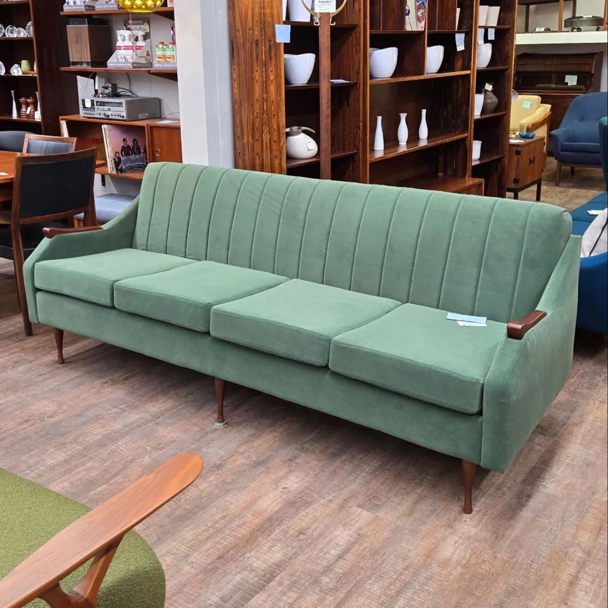 Sofa panjang gaya mid-century modern warna hijau dengan sandaran detail jahitan vertikal dan kaki kayu ramping yang estetik. Cocok untuk bagian artikel yang membahas: Mid-Century Modern atau sofa vintage.